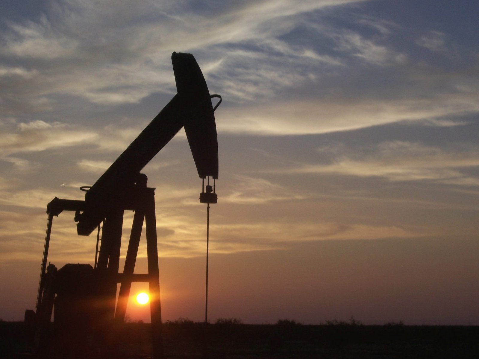 A pumping unit in silhouette against a low setting sun and streaked clouds