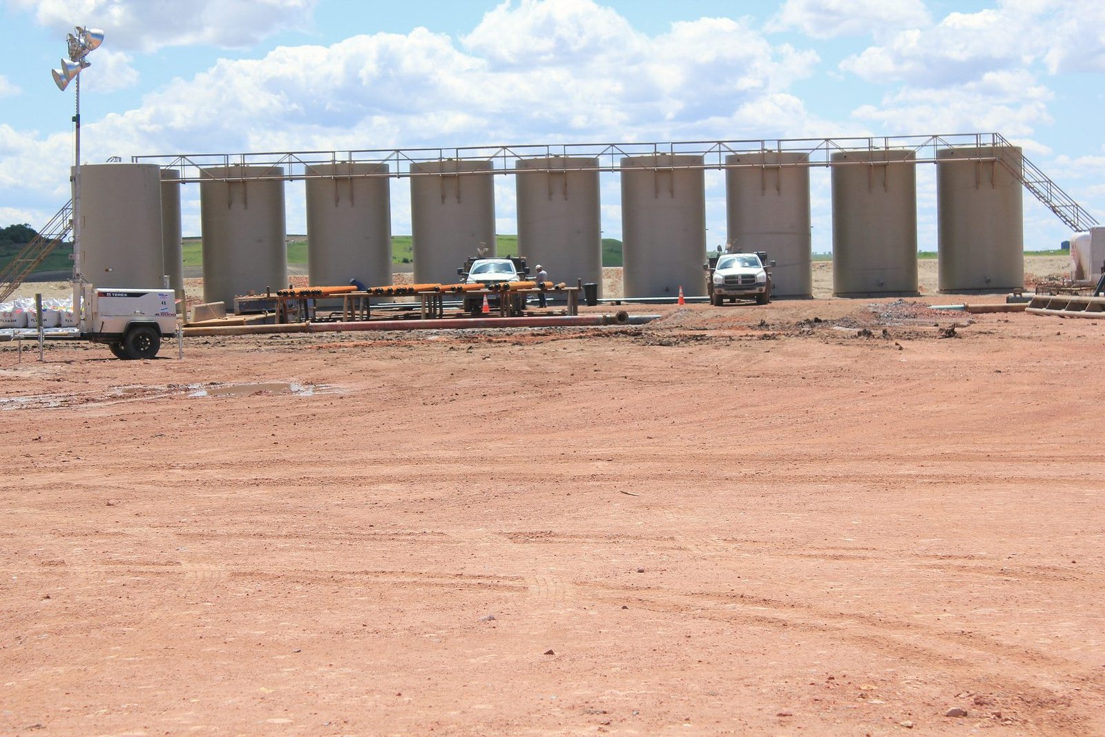 A row of tall tan storage tanks on a wide dirt pad with pickups parked in front
